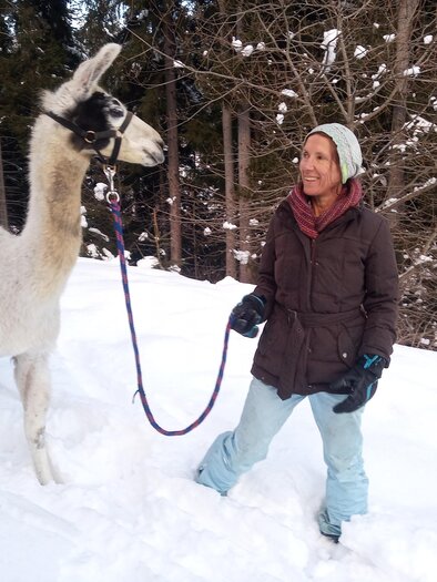 Eine Frau steht im Schnee und hält ein Lama an der Leine. Im Hintergrund sind schneebedeckte Bäume zu sehen. | © LamaTour-Orso Gigleitner
