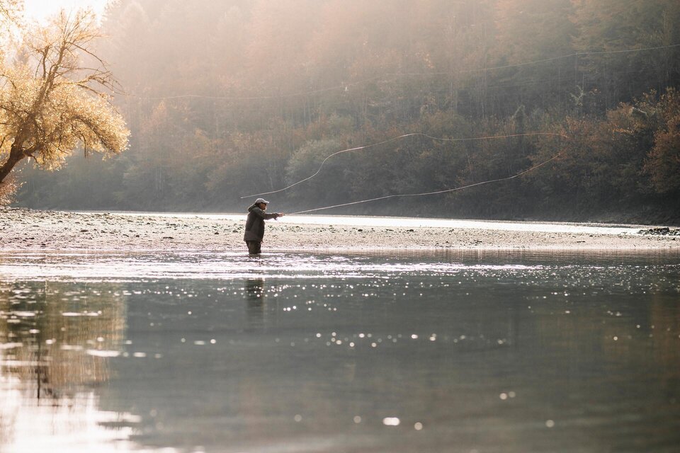 Ein Angler steht im flachen Wasser eines ruhigen Flusses. Die Umgebung ist von sanften Hügeln und Bäumen umgeben, im warmen Licht der Nachmittagssonne. | © TV Gesäuse