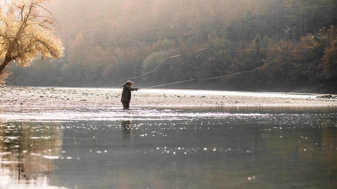 Ein Angler steht im flachen Wasser eines ruhigen Flusses. Die Umgebung ist von sanften Hügeln und Bäumen umgeben, im warmen Licht der Nachmittagssonne. | © TV Gesäuse