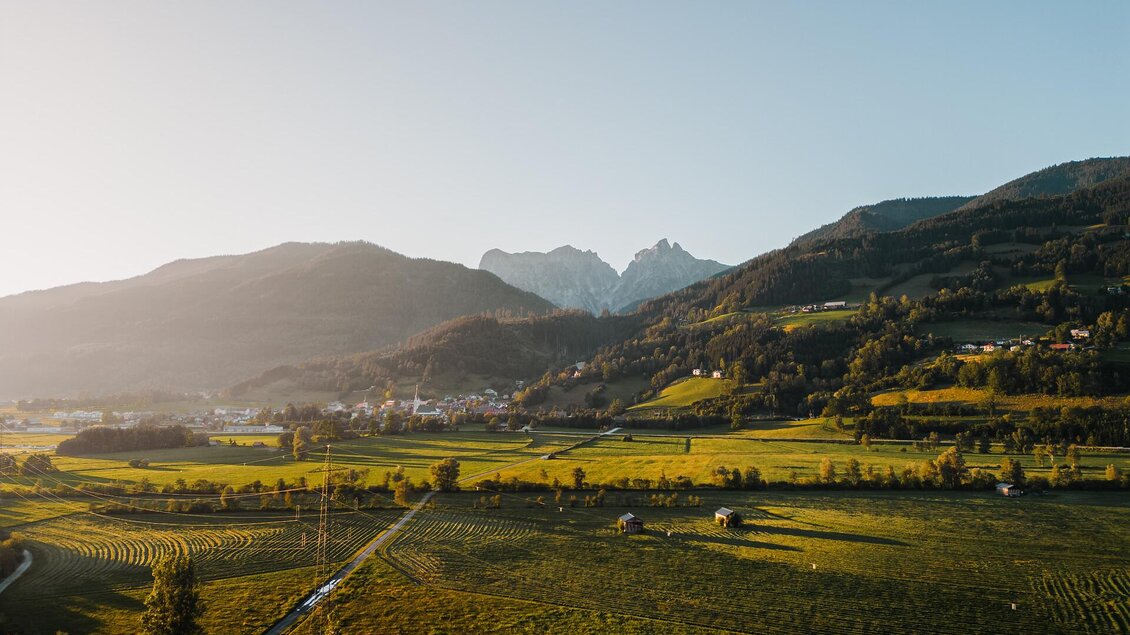 Eine malerische Landschaft mit sanften Hügeln und beeindruckenden Bergen im Hintergrund. Die Wiesen sind grün und das Licht der untergehenden Sonne verleiht der Szene einen warmen Glanz. | © TV Gesäuse