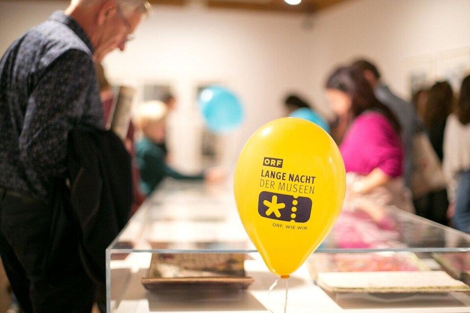 Ein gelbes Luftballon mit der Aufschrift "LANGE NACHT DER MUSEEN" steht in einem Museum. Im Hintergrund sind Besucher zu sehen, die sich die Ausstellungen ansehen. | © ORF wildbild