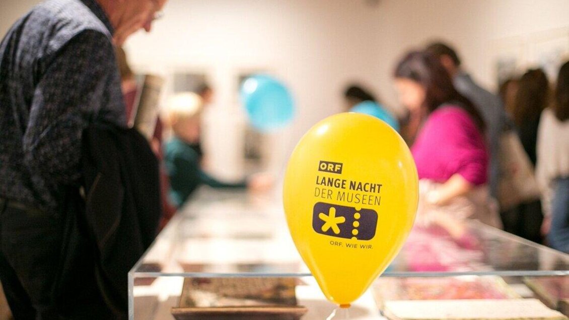 Ein gelbes Luftballon mit der Aufschrift "LANGE NACHT DER MUSEEN" steht in einem Museum. Im Hintergrund sind Besucher zu sehen, die sich die Ausstellungen ansehen. | © ORF wildbild