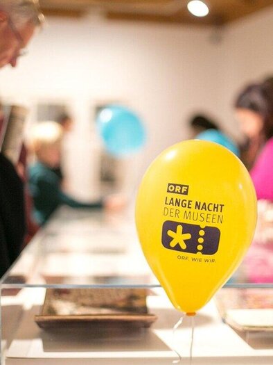 Ein gelbes Luftballon mit der Aufschrift "LANGE NACHT DER MUSEEN" steht in einem Museum. Im Hintergrund sind Besucher zu sehen, die sich die Ausstellungen ansehen. | © ORF wildbild