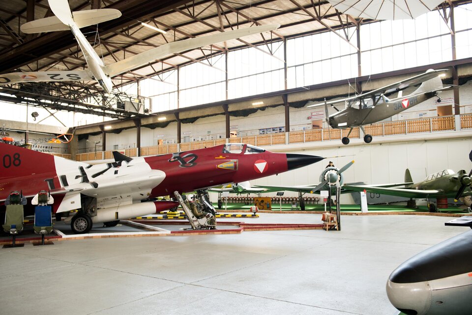 Ein Flugzeugmuseum mit verschiedenen historischen Flugzeugen. Im Vordergrund steht ein rotes Kampfflugzeug, während andere Modelle an der Decke hängen. | © Fössl Anita