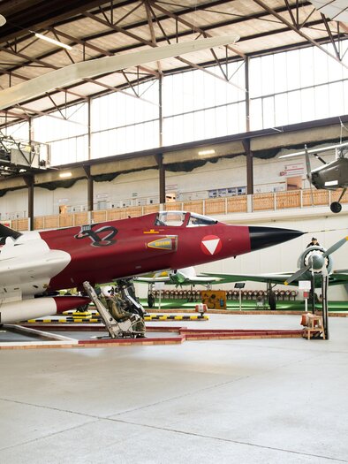 Ein Flugzeugmuseum mit verschiedenen historischen Flugzeugen. Im Vordergrund steht ein rotes Kampfflugzeug, während andere Modelle an der Decke hängen. | © Fössl Anita