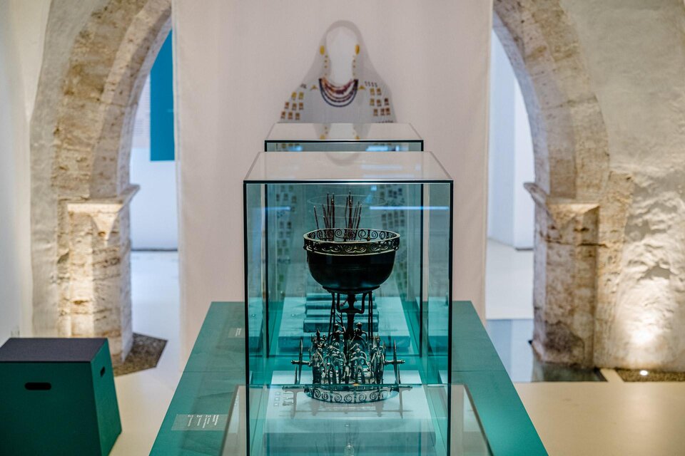 Ein elegantes Kunstwerk in einem Museum, präsentiert in einem Glasdisplay. Im Hintergrund sind historische Mauern und eine Skulptur sichtbar.