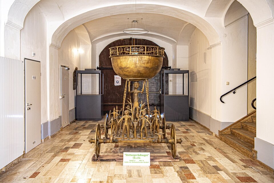 Ein elegantes Kunstwerk in einem Museum, präsentiert in einem Glasdisplay. Im Hintergrund sind historische Mauern und eine Skulptur sichtbar. | © Erlebnisregion Murtal