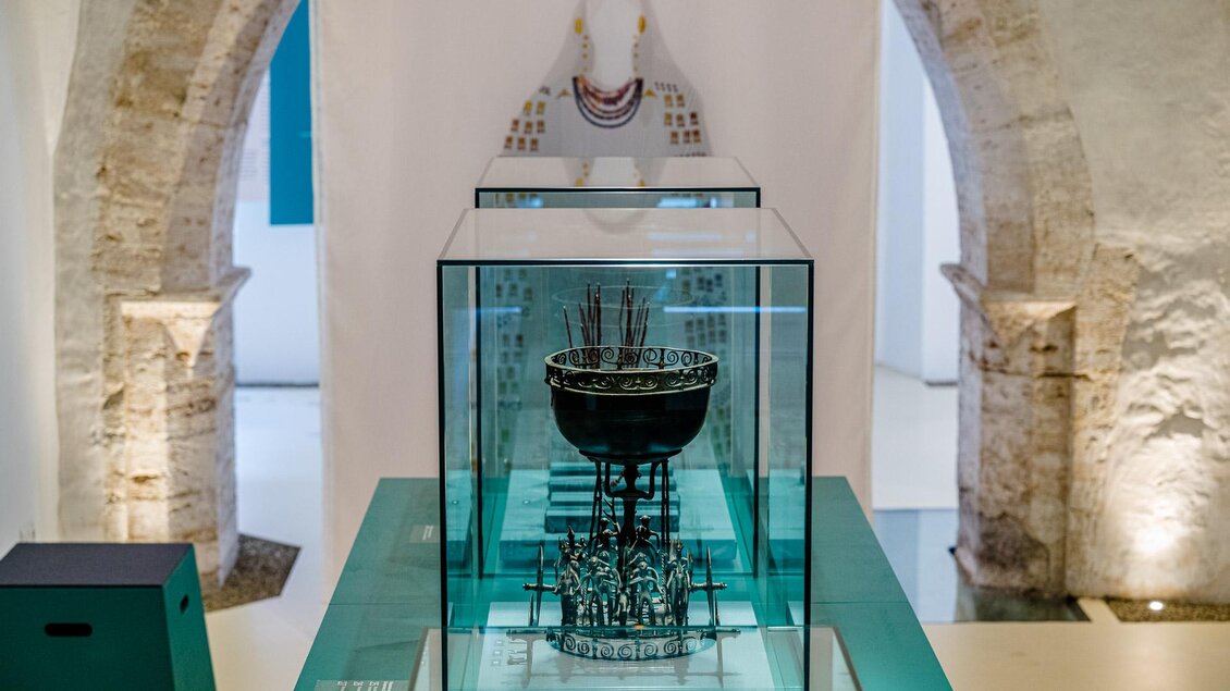 Ein elegantes Kunstwerk in einem Museum, präsentiert in einem Glasdisplay. Im Hintergrund sind historische Mauern und eine Skulptur sichtbar.