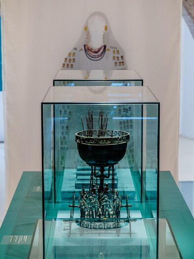 Ein elegantes Kunstwerk in einem Museum, präsentiert in einem Glasdisplay. Im Hintergrund sind historische Mauern und eine Skulptur sichtbar.