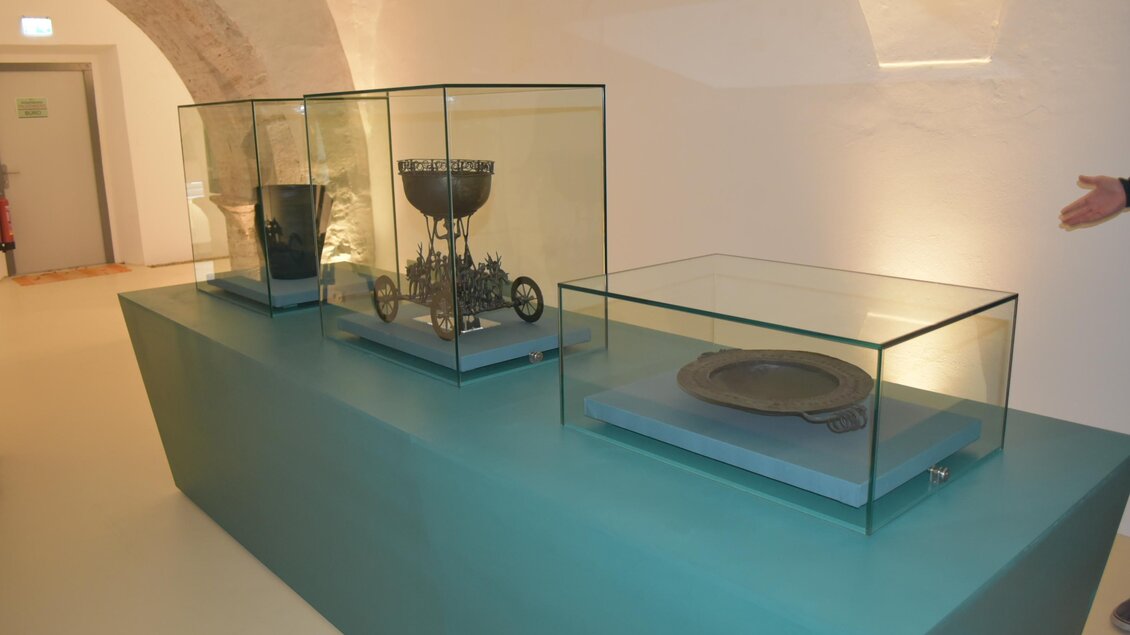 Drei Vitrinen mit antiken Objekten stehen auf einem stilvollen Tisch. Im Hintergrund ist ein heller, moderner Ausstellungsraum zu sehen.