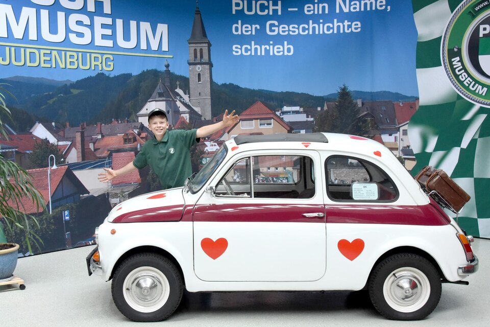 Ein Junge steht neben einem weiß-roten Auto mit Herzmotiven im PUCH Museum in Judenburg. Im Hintergrund sind die Berge und die Stadtansicht sichtbar.