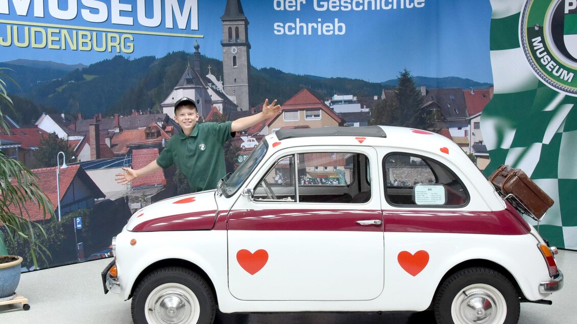 Ein Junge steht neben einem weiß-roten Auto mit Herzmotiven im PUCH Museum in Judenburg. Im Hintergrund sind die Berge und die Stadtansicht sichtbar.