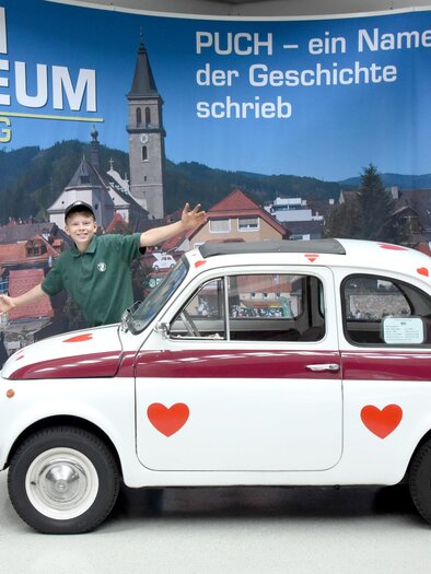 Ein Junge steht neben einem weiß-roten Auto mit Herzmotiven im PUCH Museum in Judenburg. Im Hintergrund sind die Berge und die Stadtansicht sichtbar.