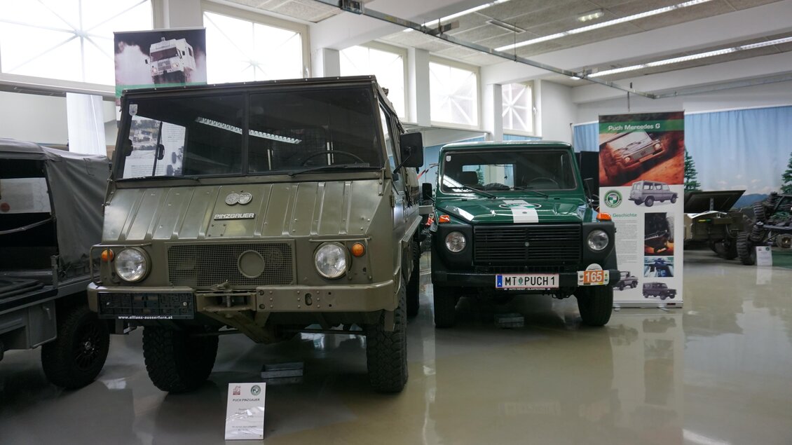 Zwei klassische Fahrzeuge stehen in einem Ausstellungsraum. Auf der linken Seite ein grüner Geländewagen und rechts ein kleiner, robuster Offroader.