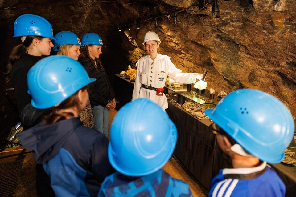 Eine Gruppe von Menschen in blauen Helmen hört einem Führer in einem Stollen zu. Der Führer zeigt verschiedene Mineralien und gibt Informationen über den Ort. | © Erlebnisregion Murtal