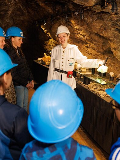 Eine Gruppe von Menschen in blauen Helmen hört einem Führer in einem Stollen zu. Der Führer zeigt verschiedene Mineralien und gibt Informationen über den Ort. | © Erlebnisregion Murtal