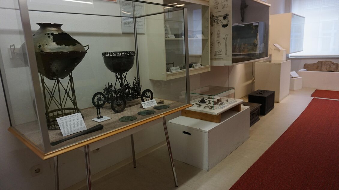 Ein Raum mit Ausstellungsstücken, darunter alte Gefäße und Maschinen. Die Objekte sind in Vitrinen präsentiert und auf einem weißen Tisch ausgestellt.