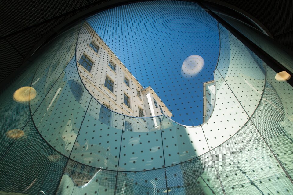 Ein Blick nach oben durch eine gläserne Struktur, die den Himmel und eine Gebäudefassade zeigt. Die Lichtspiele und die runden Formen verleihen dem Bild eine moderne Atmosphäre.