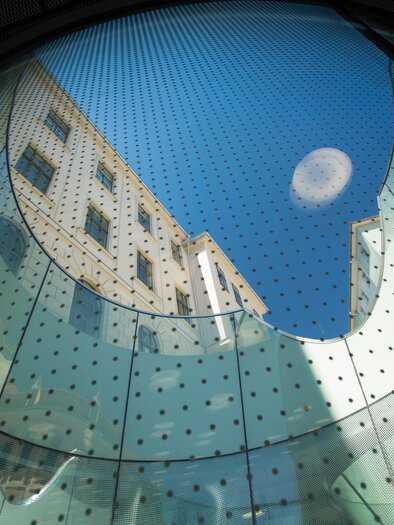 A look upwards through a glass structure that shows the sky and a building facade. The play of light and the round shapes give the image a modern atmosphere.