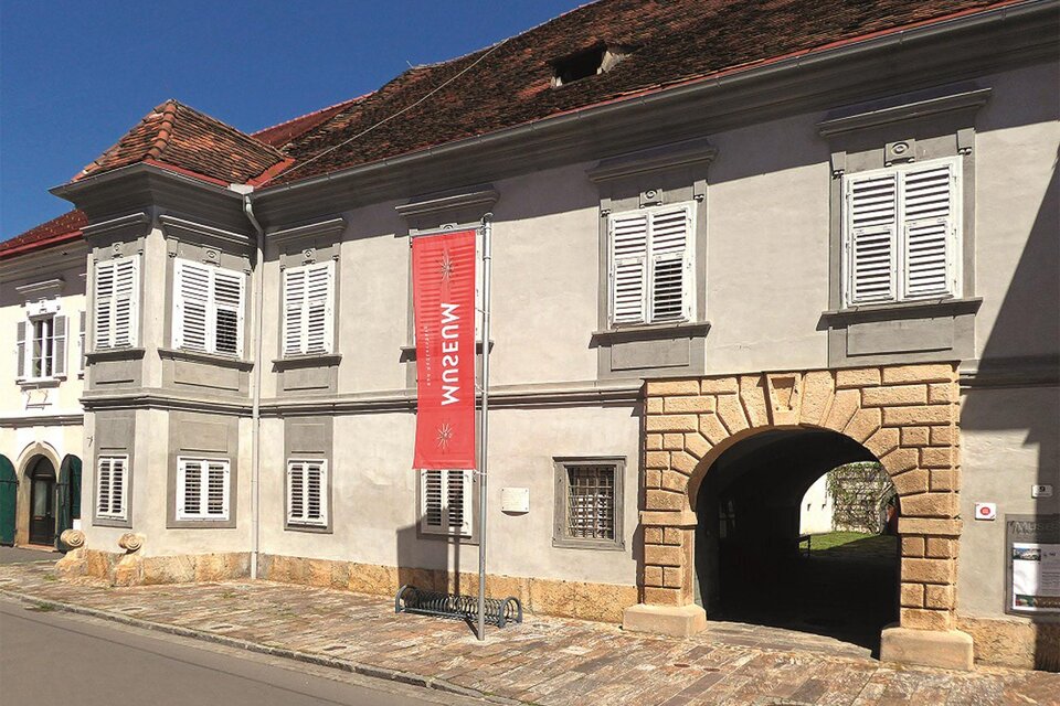 Ein historisches Gebäude mit grauer Fassade und weißen Fensterläden. Ein rotes Banner mit der Aufschrift „Museum“ hängt an der Vorderseite. | © Kranzelbinder_MiaZ