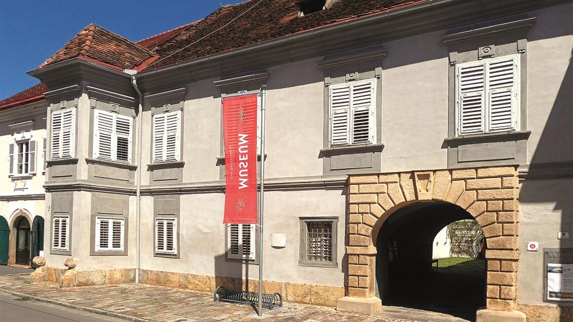 Ein historisches Gebäude mit grauer Fassade und weißen Fensterläden. Ein rotes Banner mit der Aufschrift „Museum“ hängt an der Vorderseite. | © Kranzelbinder_MiaZ