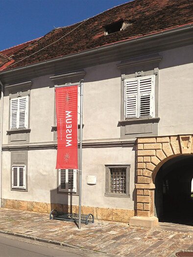 A historic building with a gray facade and white window shutters. A red banner with the inscription "Museum" hangs at the front. | © Kranzelbinder_MiaZ