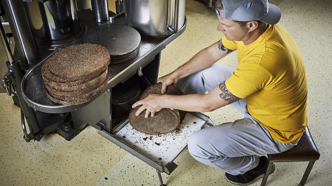 Ein Mann in einem gelben T-Shirt arbeitet an einer Maschine und hebt runde, flache Produkte. Um ihn herum liegen weitere dieser Produkte auf einem Tisch. | © Öhlmühle Fandler