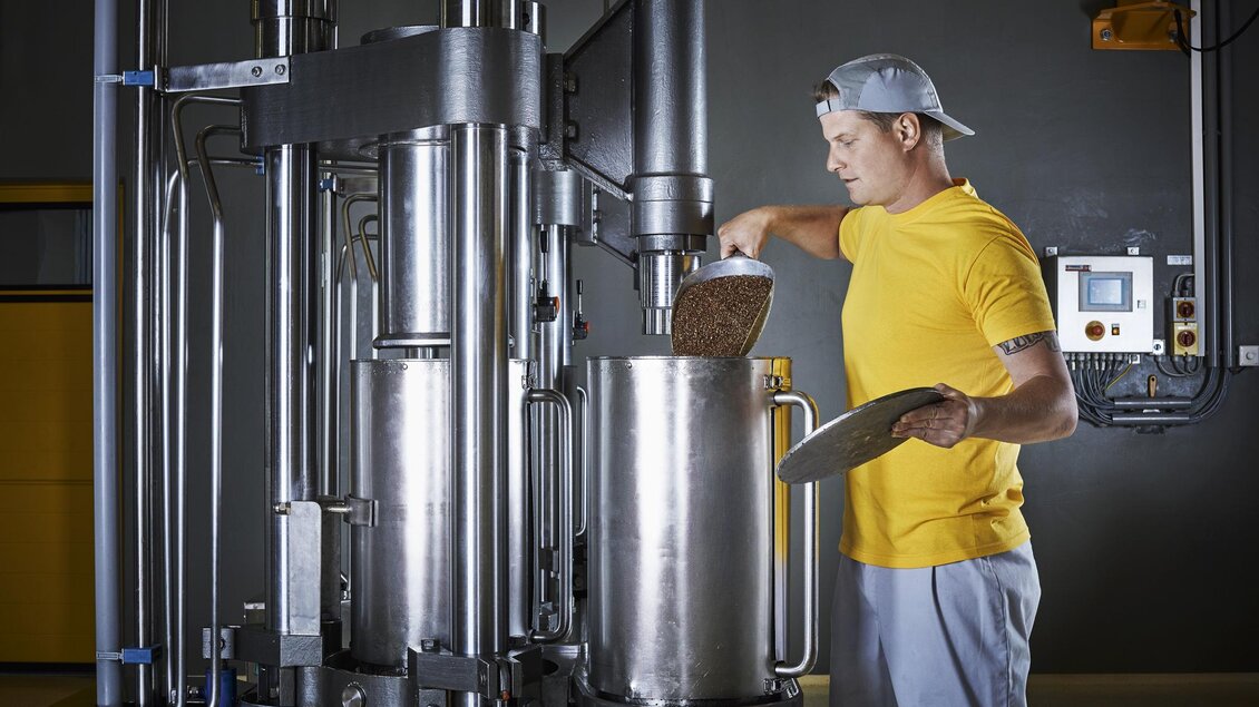 Ein Mann in einem gelben T-Shirt füllt Zutaten in eine große metallene Maschine. Die Umgebung ist eine industrielle Produktionshalle mit modernen Geräten. | © Öhlmühle Fandler