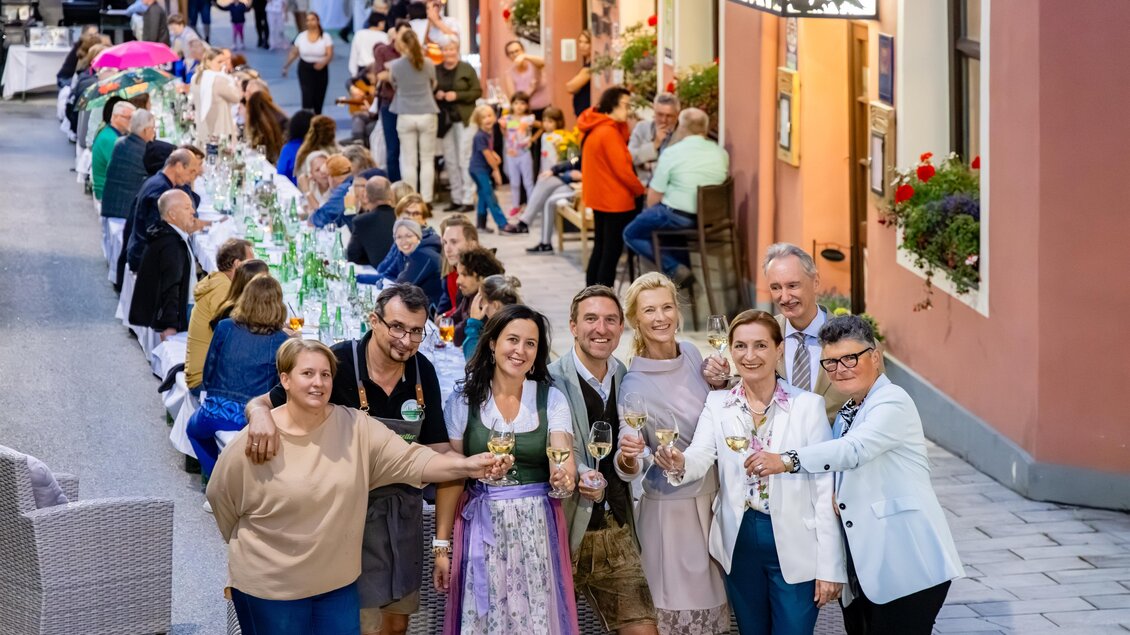 Eine festliche Straßenszene mit einer langen Tafel und Menschen, die miteinander feiern. Alle halten Gläser und genießen die gesellige Atmosphäre. | © Fred Lindmoser | ARGE Genussmeile Wiener Straße