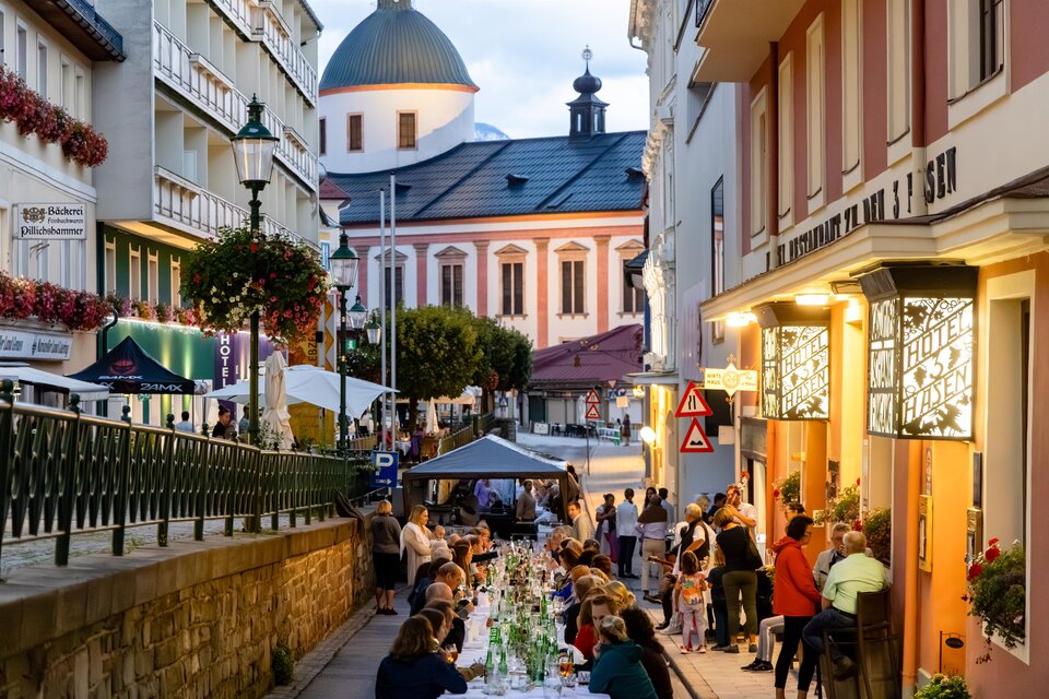 Eine belebte Straße mit einem langen Tisch, an dem Menschen essen und trinken. Im Hintergrund ist eine hübsche Kirche zu sehen, während die Abendbeleuchtung eine gemütliche Atmosphäre schafft. | © Fred Lindmoser | ARGE Genussmeile Wiener Straße
