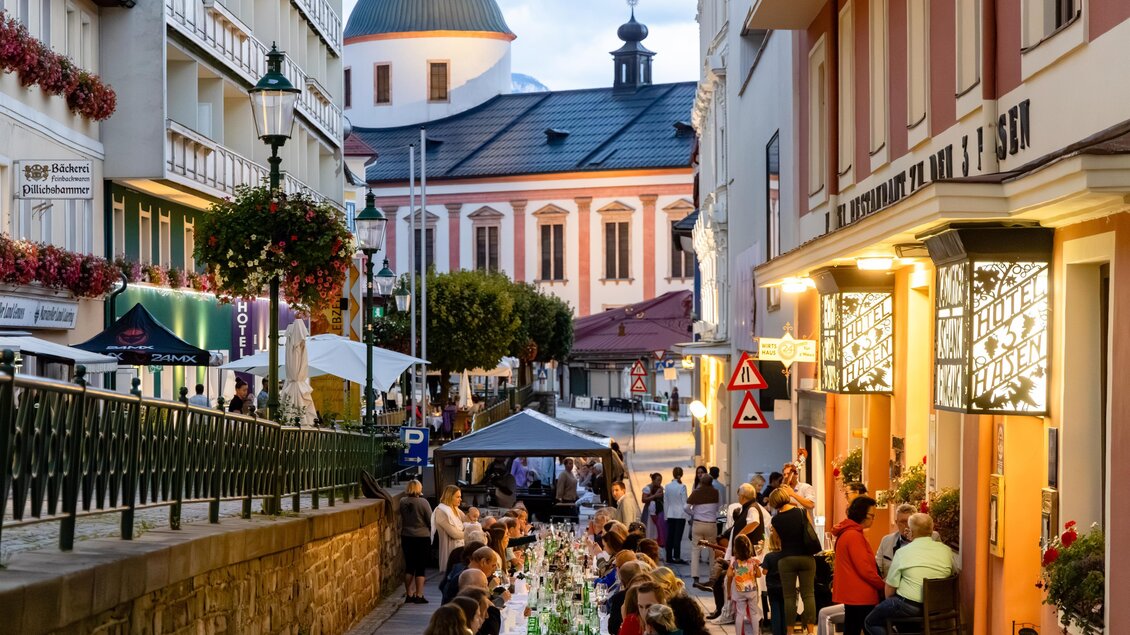 Eine belebte Straße mit einem langen Tisch, an dem Menschen essen und trinken. Im Hintergrund ist eine hübsche Kirche zu sehen, während die Abendbeleuchtung eine gemütliche Atmosphäre schafft. | © Fred Lindmoser | ARGE Genussmeile Wiener Straße