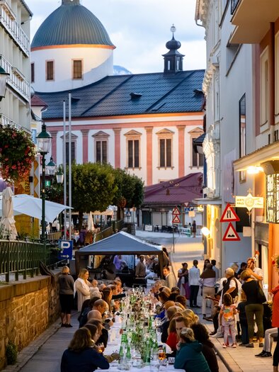 Eine belebte Straße mit einem langen Tisch, an dem Menschen essen und trinken. Im Hintergrund ist eine hübsche Kirche zu sehen, während die Abendbeleuchtung eine gemütliche Atmosphäre schafft. | © Fred Lindmoser | ARGE Genussmeile Wiener Straße