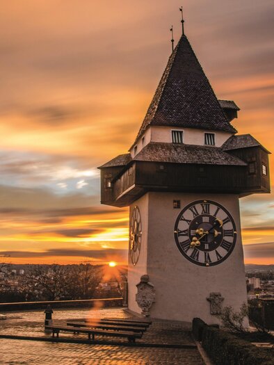 A beautiful view of an old tower at sunset. The sky is tinted in warm colors and the city is visible in the background. | © Graz Tourismus - Markus Spenger