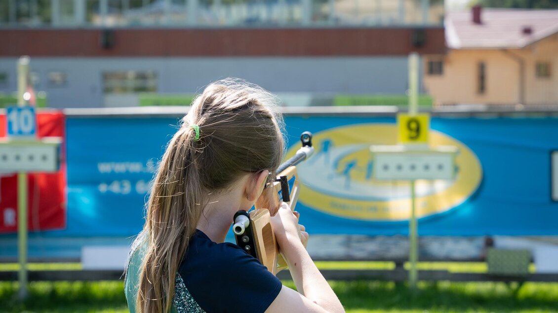 Ein Mädchen zielt mit einem Luftgewehr auf eine Zielscheibe im Freien. Im Hintergrund ist eine alpine Landschaft zu sehen. | © TVB/Laura Passenbrunner