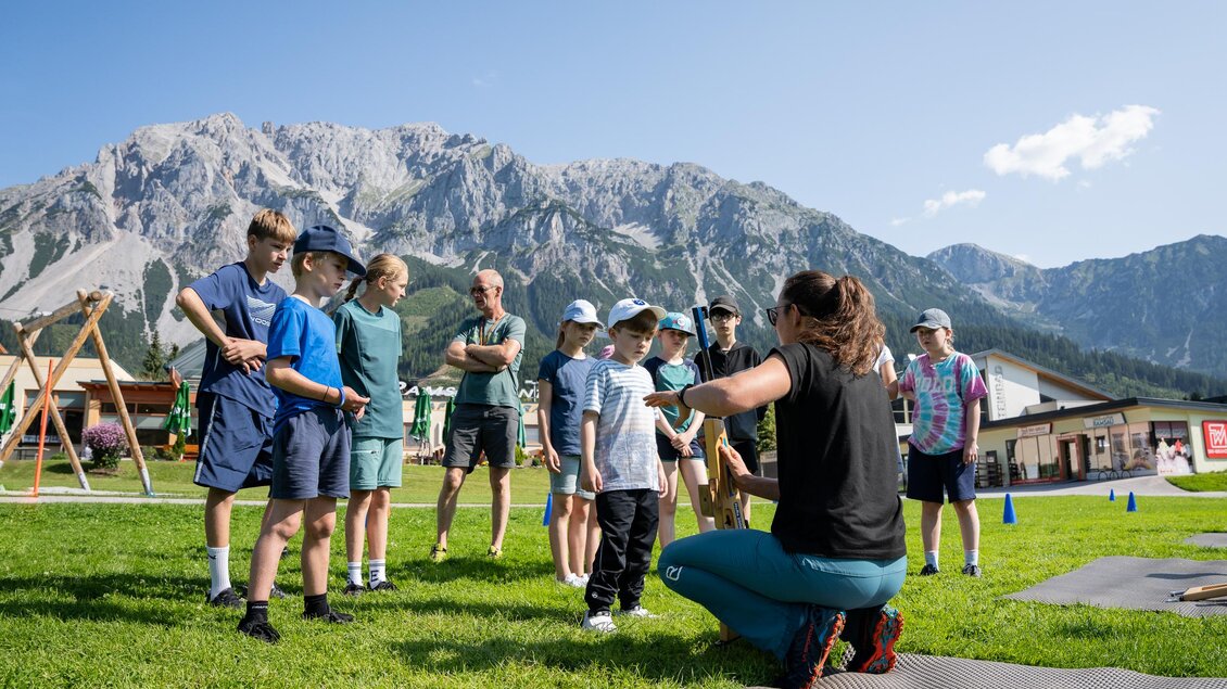 Eine Gruppe von Kindern lauscht aufmerksam einer Trainerin im Freien. Im Hintergrund sind Berge und Spielgeräte zu sehen. | © TVB/Laura Passenbrunner