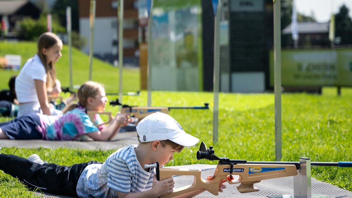 Eine Gruppe von Kindern liegt im Gras und übt Schießen mit Sportgewehren. Im Hintergrund sind Berge und eine Schießstandanlage zu sehen. | © TVB/Laura Passenbrunner
