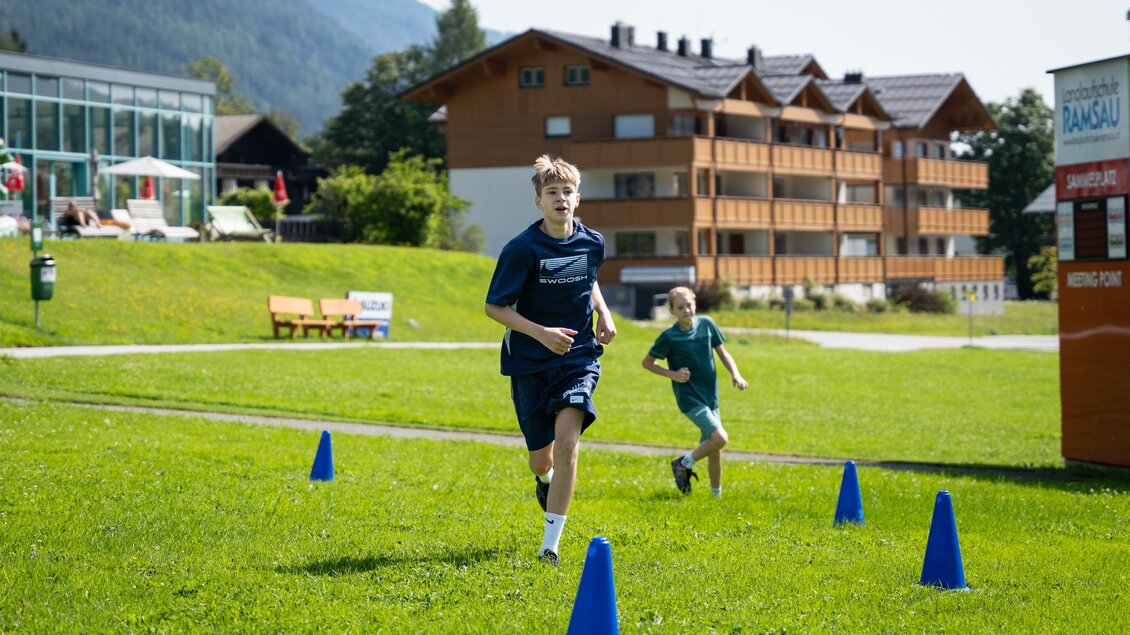 Zwei Jungen laufen auf einer Wiese zwischen blauen Hütchen. Im Hintergrund sind Gebäude und Bäume sichtbar. | © TVB/Laura Passenbrunner
