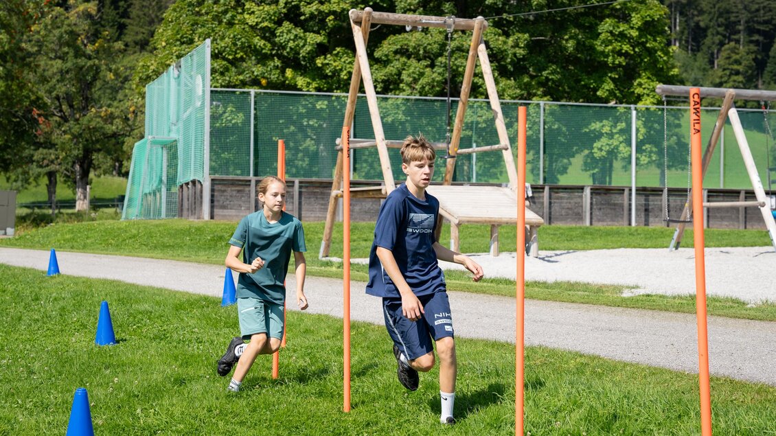Zwei Kinder laufen auf einem Sportplatz zwischen Trainingshindernissen. Im Hintergrund sind Bäume und ein Tennisplatz zu sehen. | © TVB/Laura Passenbrunner