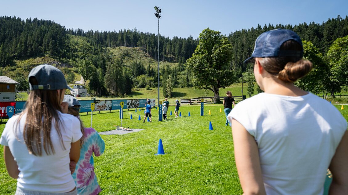 Eine Gruppe von Kindern spielt auf einem grünen Sportplatz mit blauen Hütchen. Im Hintergrund sind Bäume und Hügel zu sehen. | © TVB/Laura Passenbrunner