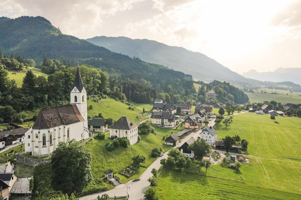 Eine malerische Landschaft mit einer Kirche und kleinen Häusern. Im Hintergrund sind grüne Hügel und Berge zu sehen.