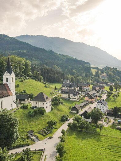 Eine malerische Landschaft mit einer Kirche und kleinen Häusern. Im Hintergrund sind grüne Hügel und Berge zu sehen.
