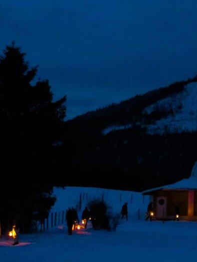 Eine ruhige Winterlandschaft bei Nacht mit einem beleuchteten Gebäude und schneebedeckten Bergen im Hintergrund. Fackeln säumen den Weg und schaffen eine gemütliche Atmosphäre. | © Anna Scherfler