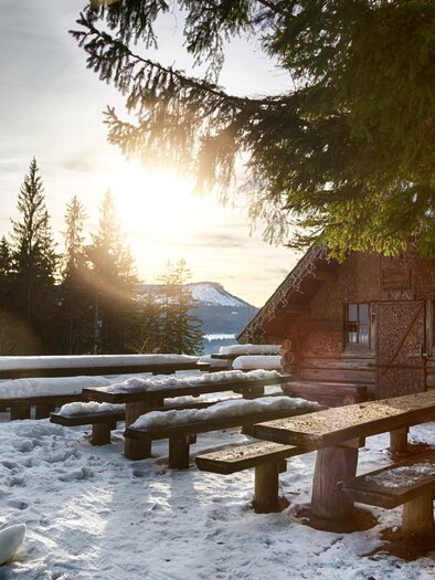 Eine gemütliche Hütte in einer verschneiten Landschaft. Im Vordergrund stehen Holzbänke und der Sonnenuntergang strahlt warmes Licht aus. | © Fred Lindmoser