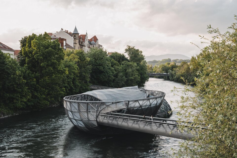Eine moderne Brücke über einen Fluss, umgeben von Bäumen und historischen Gebäuden. Der Himmel ist bewölkt und die Landschaft wirkt ruhig und einladend. | © Region Graz_HOUSE of MANAO