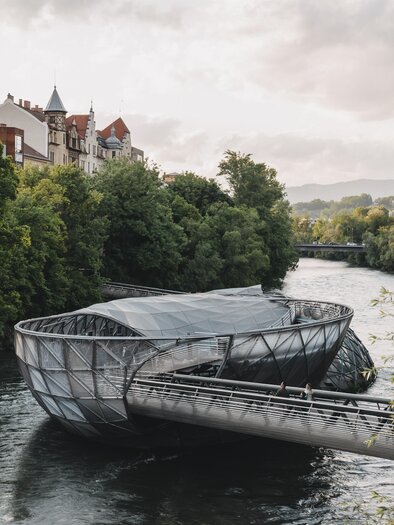 A modern bridge over a river, surrounded by trees and historical buildings. The sky is cloudy and the landscape looks peaceful and inviting. | © Region Graz_HOUSE of MANAO