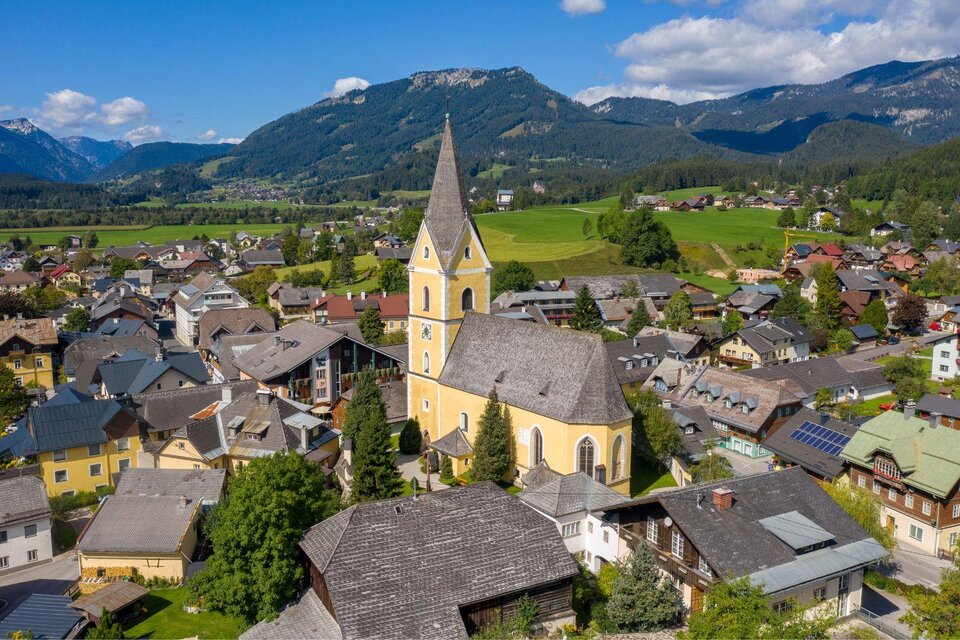 Guter Blick auf die gelbe Kirche und das Ortszentrum von Bad Mitterndorf an einem schönen Tag mit blauem, nahezu wolkenlosem Himmel. Die umliegenden Wiesen und Wälder zeigen sich in sattem Grün, während die Berge im Hintergrund gut zu erkennen sind. | © Karl Grieshofer