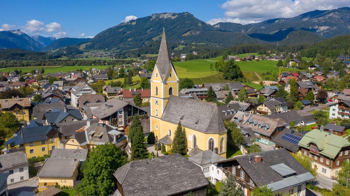 Guter Blick auf die gelbe Kirche und das Ortszentrum von Bad Mitterndorf an einem schönen Tag mit blauem, nahezu wolkenlosem Himmel. Die umliegenden Wiesen und Wälder zeigen sich in sattem Grün, während die Berge im Hintergrund gut zu erkennen sind. | © Karl Grieshofer