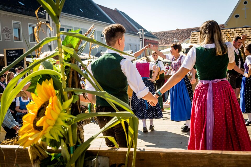 Eine fröhliche Tänzergruppe in traditionellen Trachten tanzt zusammen auf einem Fest. Im Vordergrund sind Sonnenblumen und eine festliche Atmosphäre sichtbar. | © TV Südsteiermark stephan friesinger