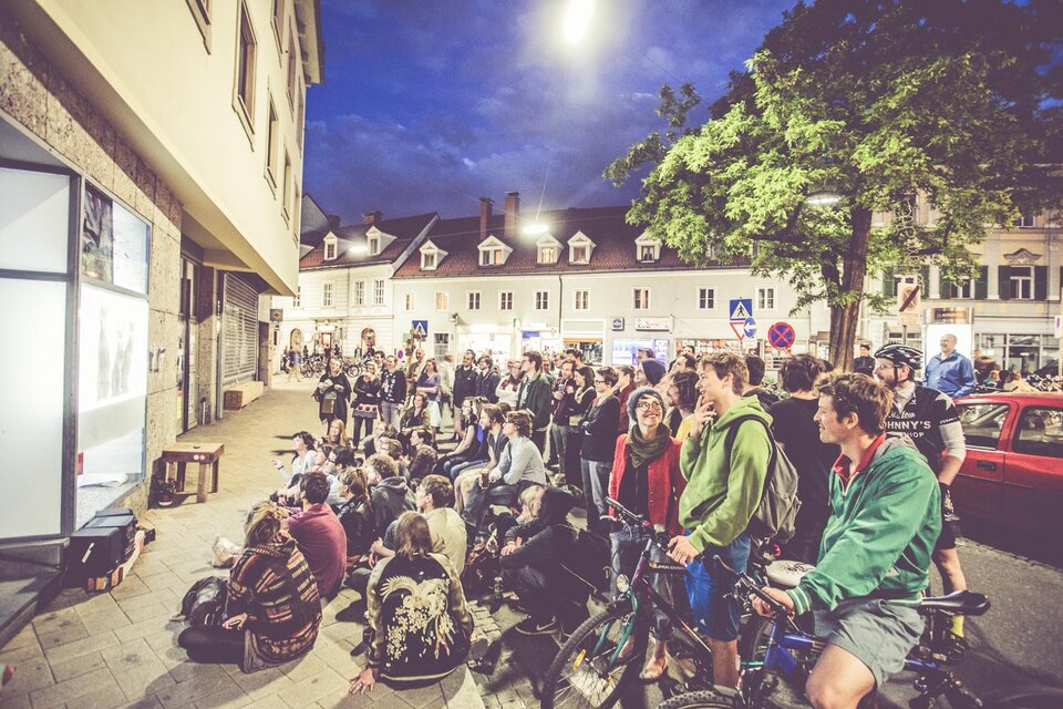 Eine große Menschenmenge sitzt und steht vor einem Gebäude in der Stadt. Es ist Abend und eine Vorstellung wird an einer Wand gezeigt. | © Lupi Spuma
