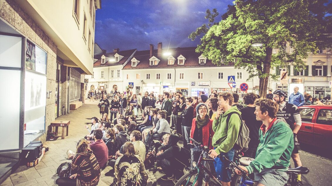 Eine große Menschenmenge sitzt und steht vor einem Gebäude in der Stadt. Es ist Abend und eine Vorstellung wird an einer Wand gezeigt. | © Lupi Spuma